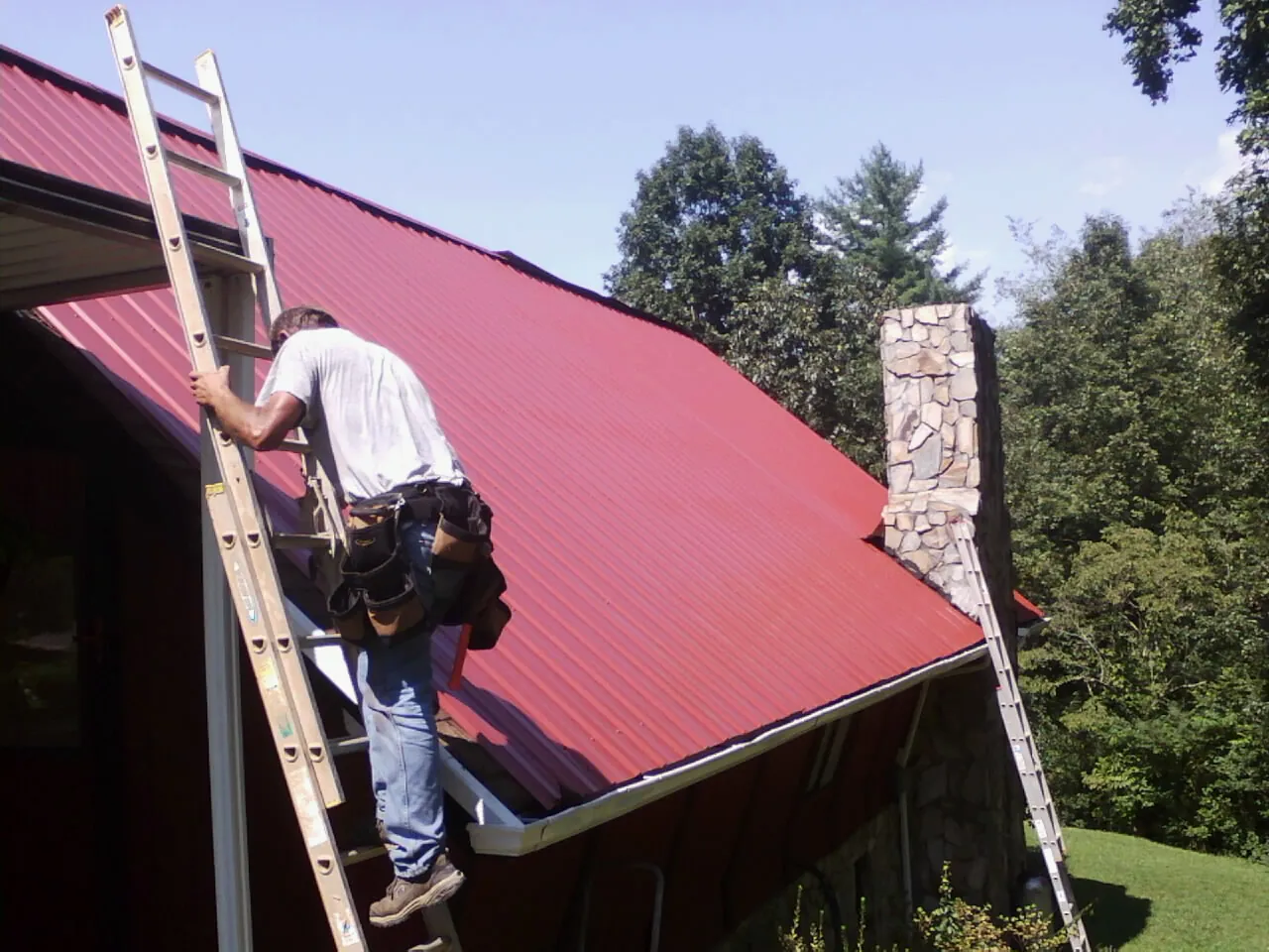 Roofer on a ladder beside a red metal roof and stone chimney — Soffit & Fascia Repair by Roofing Brothers in Mineral